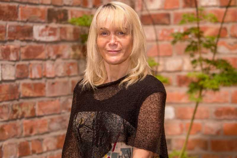 blonde woman in a mesh black top, standing against a red brick background